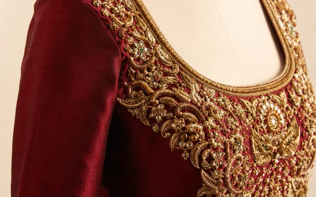 Deep wine-red raw-silk bridal blouse on a mannequin with heavy raised gold zardosi and kundan embroidery across the neckline and yoke, plain sleeve alongside — Akira Fashion Studio Bangalore