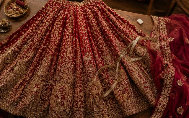Deep-red bridal lehenga skirt with gold zari and maggam embroidery spread on a wooden studio cutting table with measuring tape and matching dupatta — Akira Fashion Studio Bangalore