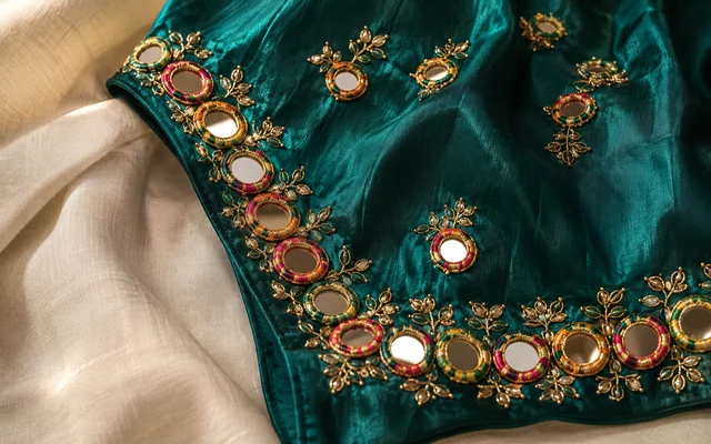 Deep emerald festive blouse with hand-set round mirror (sheesha) work caged in colourful thread and gold aari accents on ivory linen — Akira Fashion Studio Bangalore